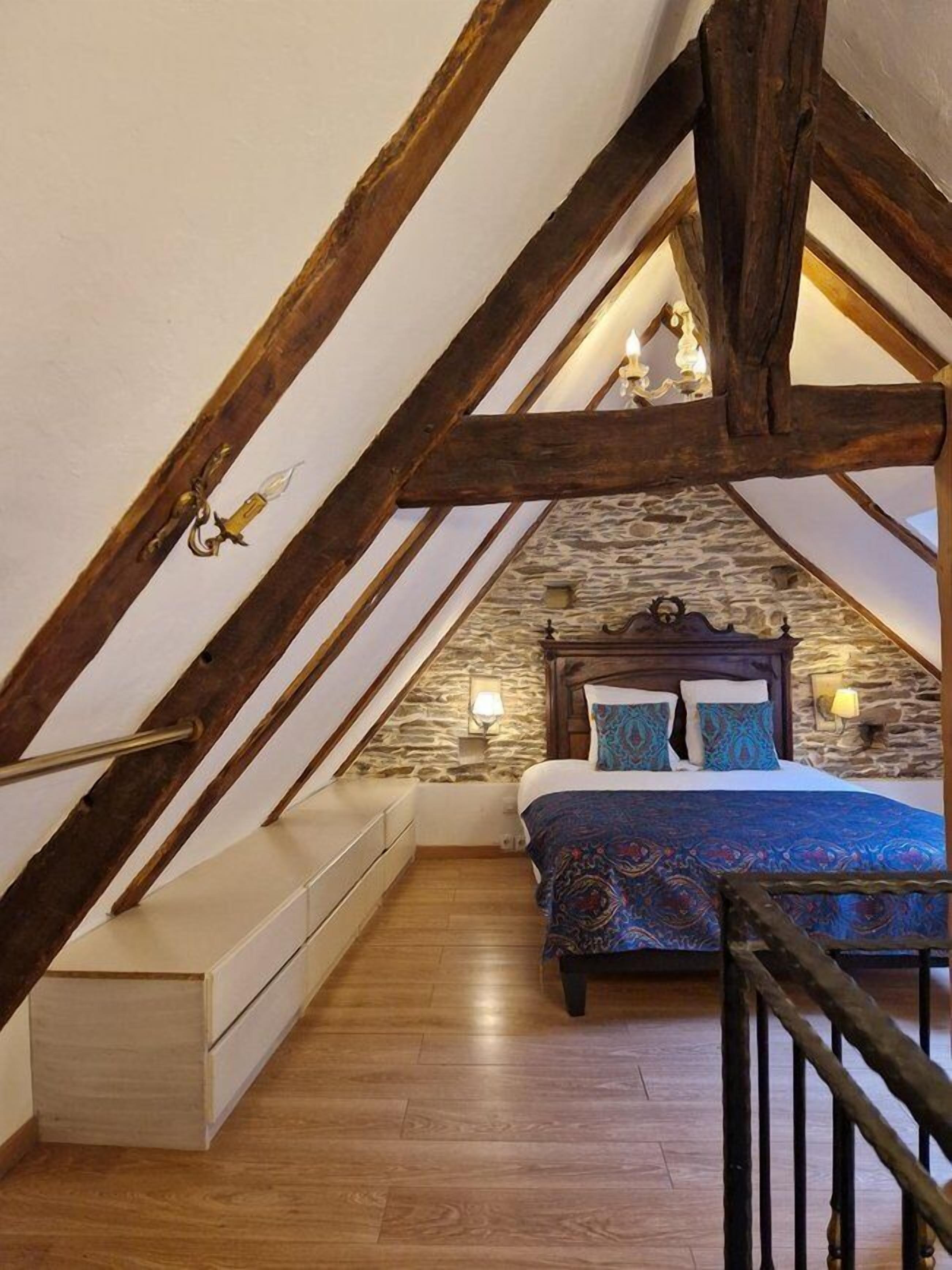 A bedroom with a sloped ceiling, stone wall, wooden beams, a bed with patterned bedding, bedside lamps, and a built-in bench.