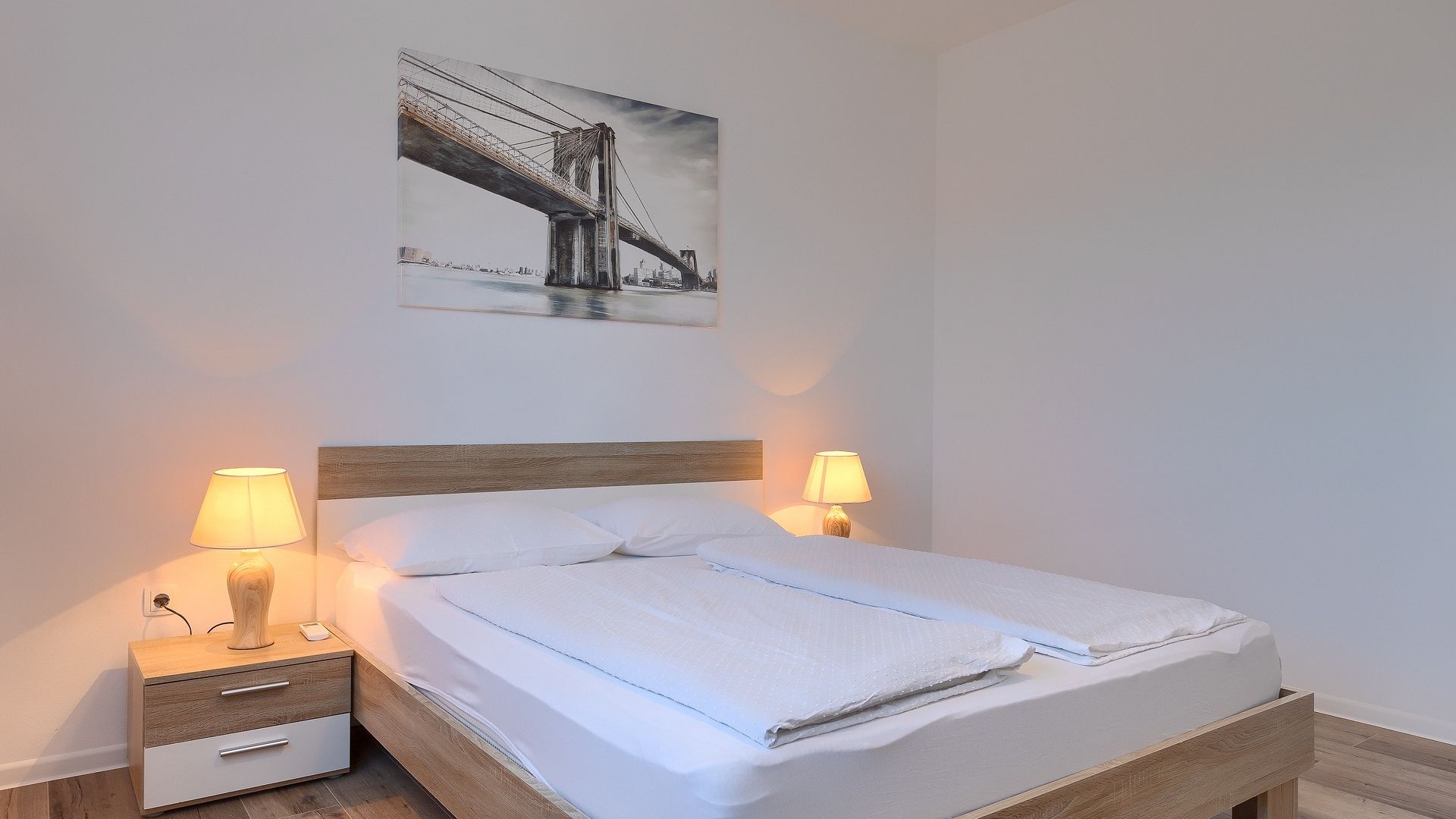 A neatly made bed with a wooden headboard, two bedside lamps, a small drawer, and a framed picture of a bridge on the wall.