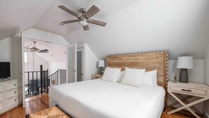 A bedroom with a large bed, two bedside tables with lamps, a ceiling fan, a wooden bench, and a staircase leading to an upper floor. - Cozy 2BR Carriage in the heart of Downtown! (Charleston)