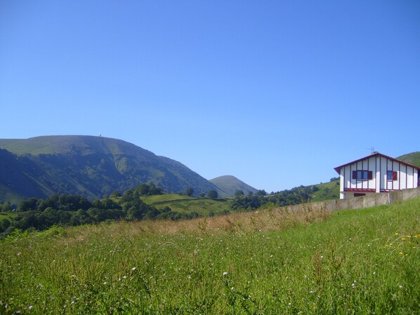 Paisagem rural com um pequeno edifício numa encosta, rodeado por campos verdes e colinas.