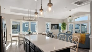 A modern kitchen with a central island, dining area with chairs, and a living area with a sofa and plant.