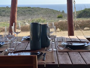 A wooden table set for dining with wine glasses, plates, and a pitcher, overlooking a coastal landscape. - Experience an adventurous way of holiday? Quinta Aurora, a life time experience. (Burgau)