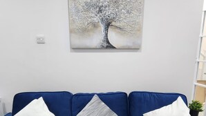 Un canapé bleu avec trois coussins blancs, un tableau représentant un arbre accroché au mur et une prise de courant sur le mur.