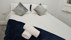 Un lit bien fait, recouvert d’une couverture bleu foncé et agrémenté de deux oreillers, un blanc et un gris, dans une pièce simple aux murs blancs et dotée d’une fenêtre.