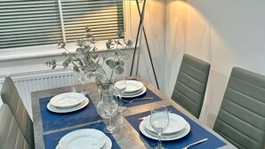 Une table à manger dressée avec des assiettes, des verres et des couverts, entourée de chaises, avec une lampe sur pied et une fenêtre avec des stores en arrière-plan.