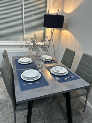 A dining table set with plates, glasses, and cutlery, flanked by chairs, in a room with a radiator and a lamp. - House Sleeps 6 With Free Parking (Staines)