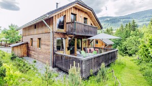 Une cabane en bois dotée d’un balcon, entourée d’une végétation luxuriante et avec une chaîne de montagnes en toile de fond.