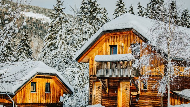Une cabane en bois dotée d’un balcon et d’un toit enneigé, entourée d’arbres et surplombant un paysage montagneux.