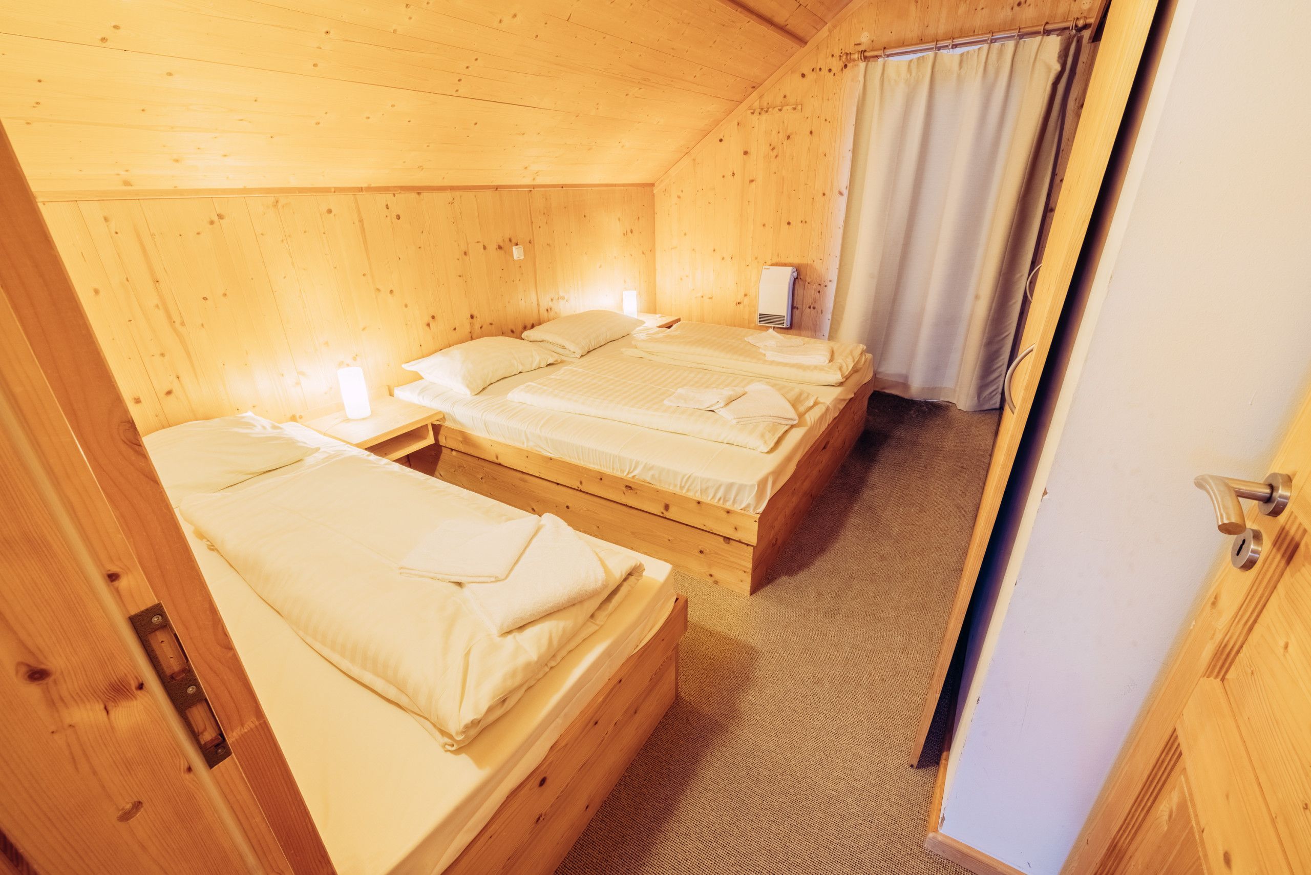 A wooden cabin with three single beds, each with a white mattress and pillows, and a small bedside table with a lamp.