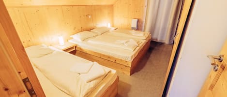 A wooden cabin with three single beds, each with a white mattress and pillows, and a small bedside table with a lamp.