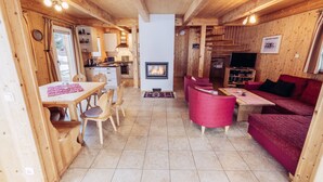 A cozy wooden cabin interior with a dining area, a fireplace, a living room with a red sofa, and a kitchen.