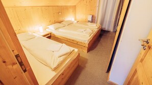 A wooden cabin with three single beds, each with a white mattress and pillows, and a small bedside table with a lamp.