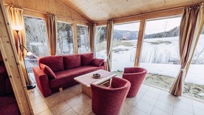 A cozy room with a red sofa, a wooden coffee table, and two red chairs. Large windows offer a view of a snowy landscape.