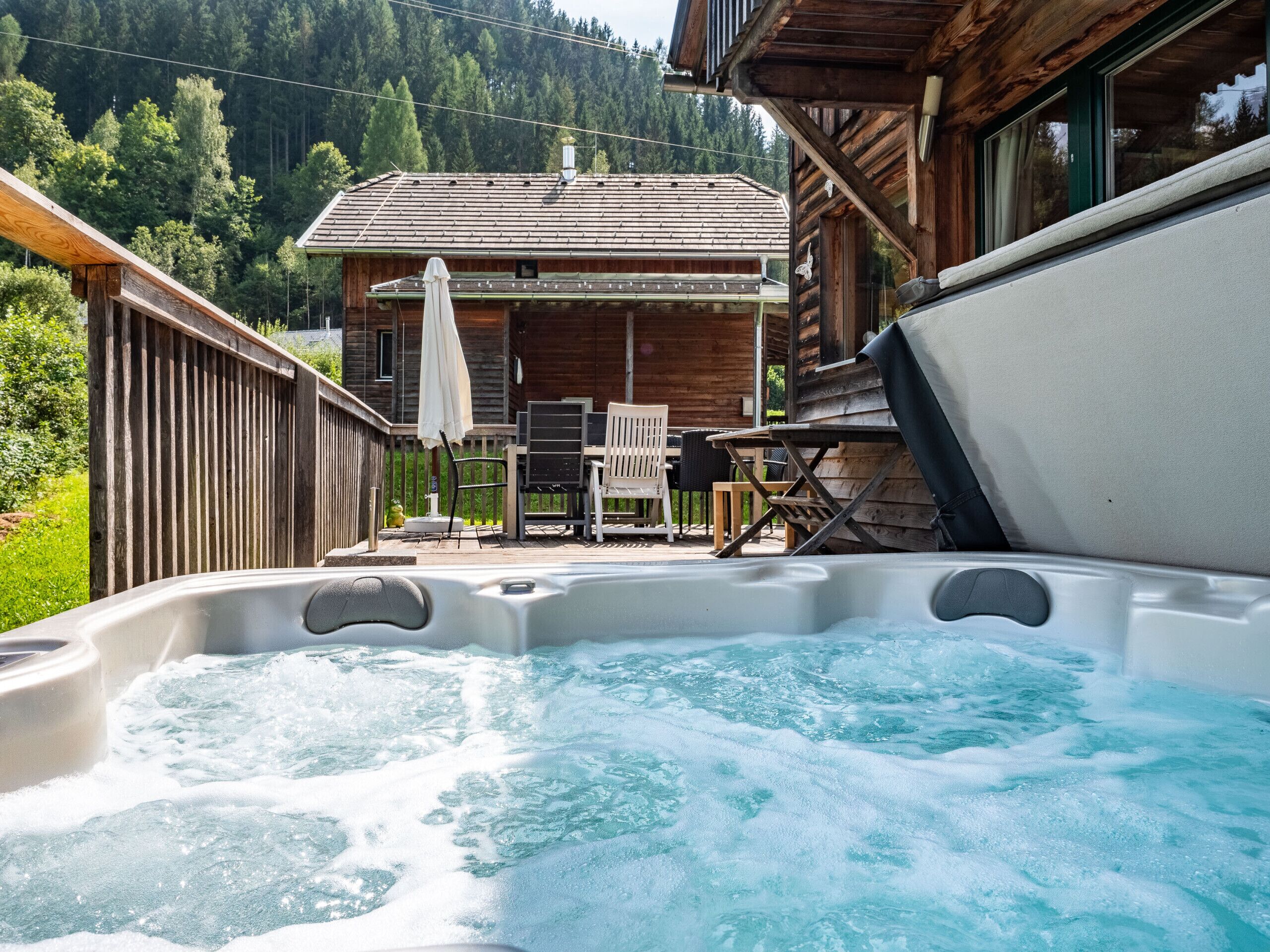Una vasca idromassaggio con un pontile in legno e mobili da esterno, circondata da una recinzione di legno con una montagna sullo sfondo.