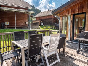 Un patio in legno con tavolo e sedie, un barbecue e vista su una baita dalla facciata in legno.