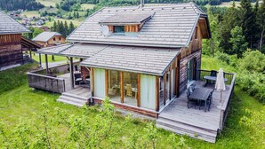 A wooden cabin with a glass-enclosed patio, surrounded by lush greenery and a mountainous landscape. - Woody 12 - with sauna & 2 cozy terraces (St. Georgen am Kreischberg)