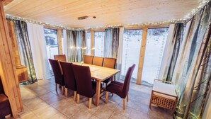 Um quarto com painéis de madeira, uma mesa de jantar e cadeiras, janelas amplas com cortinas, e um pequeno banco de madeira.