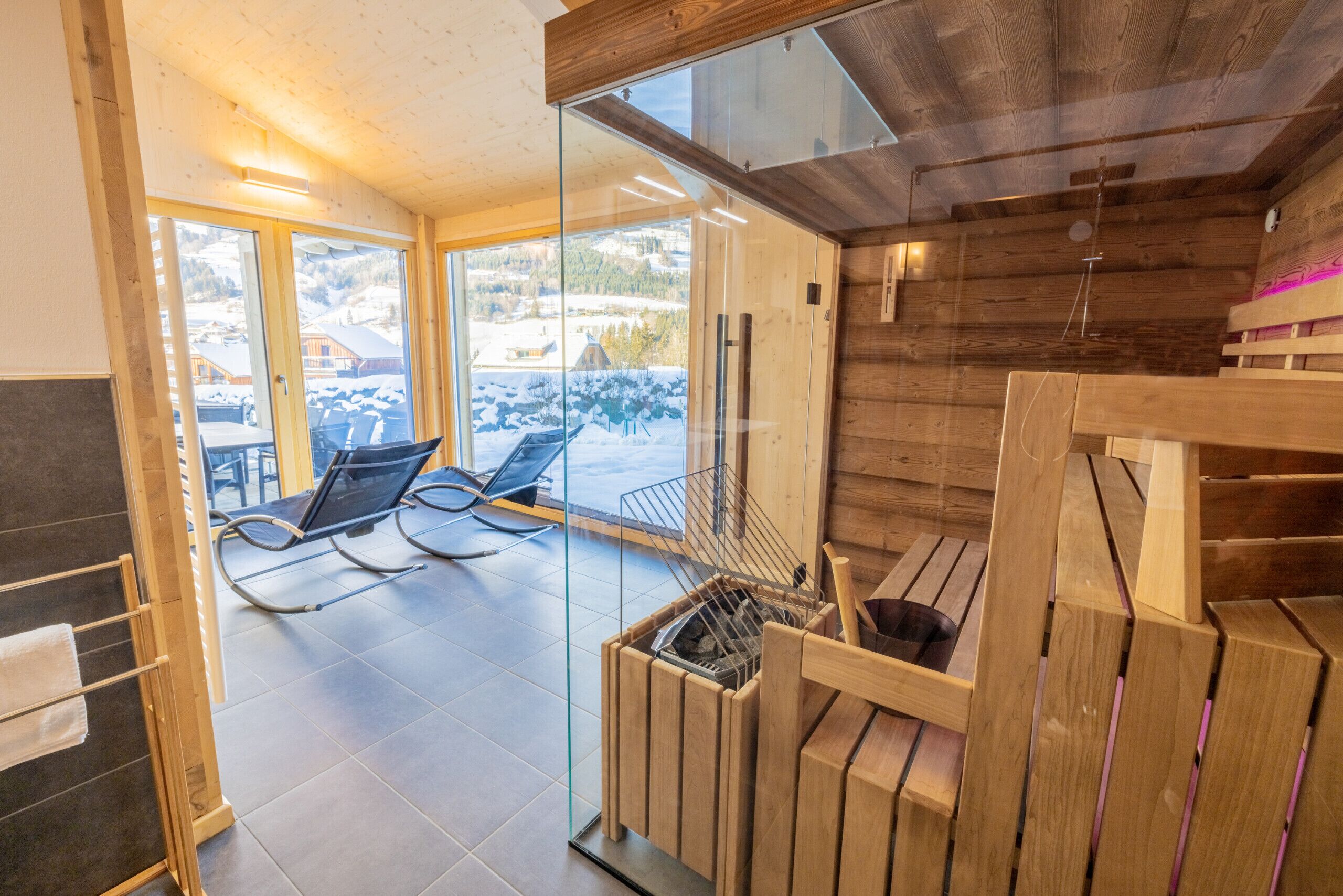 A modern wooden interior with a glass door leading to a balcony with outdoor seating and a view of a snowy landscape.