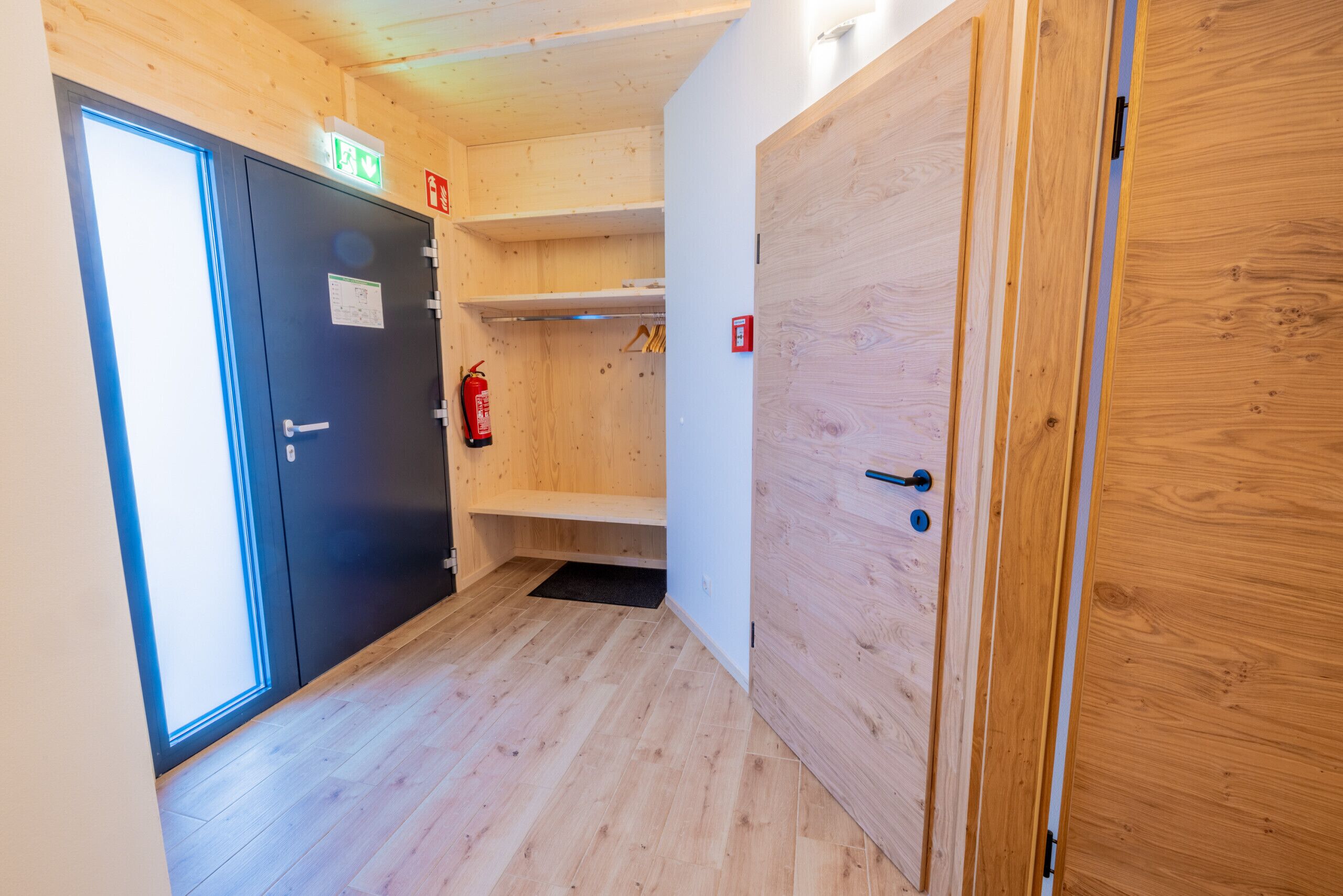 A narrow hallway with wooden walls and floors, a black door with an emergency exit sign, and a fire extinguisher mounted on the wall.