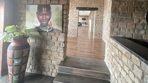A stone-walled corridor with a wooden staircase leading to a room. A large framed picture is mounted on the wall, and a potted plant is visible on the left. - Katurum Lodge Kidepo  (Loitanit)