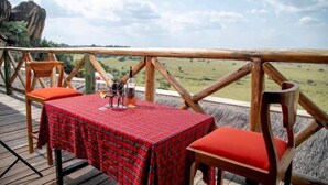 A table with a plaid tablecloth and two chairs with red cushions on an outdoor deck with a wooden railing, overlooking a grassy landscape. - Katurum Lodge Kidepo  (Loitanit)