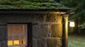A stone cottage with a moss-covered roof, a lit window, and a stone wall. - The Dell of Abernethy (Nethy Bridge)