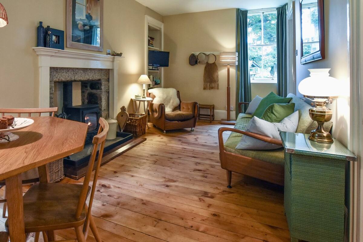 A cozy living room with a fireplace, wooden flooring, a dining table with chairs, a sofa, and a TV mounted on the wall.