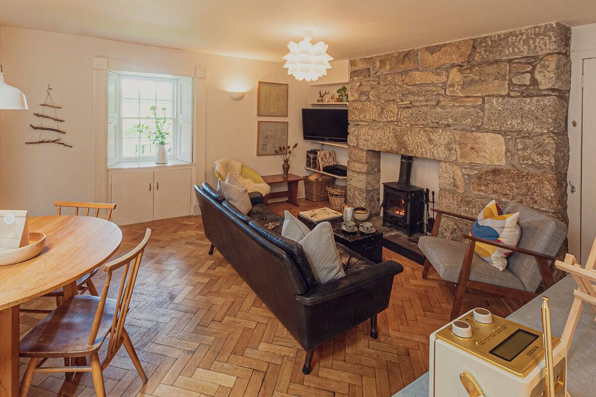 A cozy living room with a stone fireplace, a black sofa, wooden flooring, and a dining area with a table and chairs.