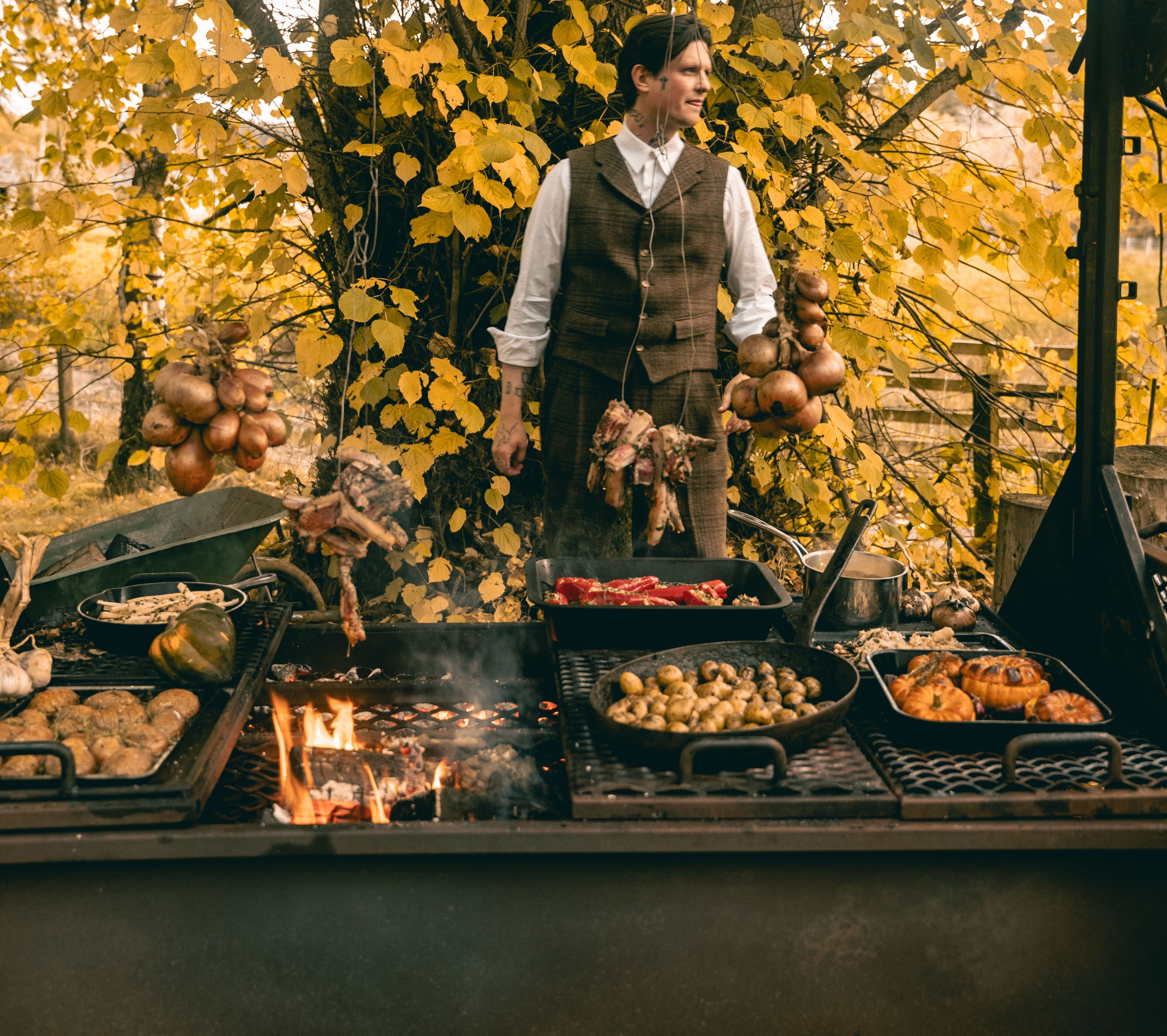 A person grilling various foods outdoors with a fire pit and a grill in the foreground.