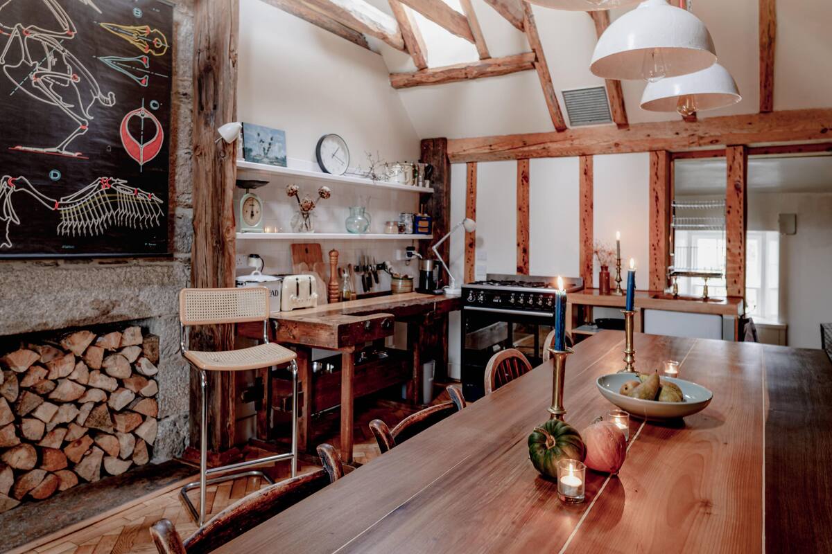 A rustic kitchen with wooden beams, a stone fireplace, and a wooden dining table with candles.