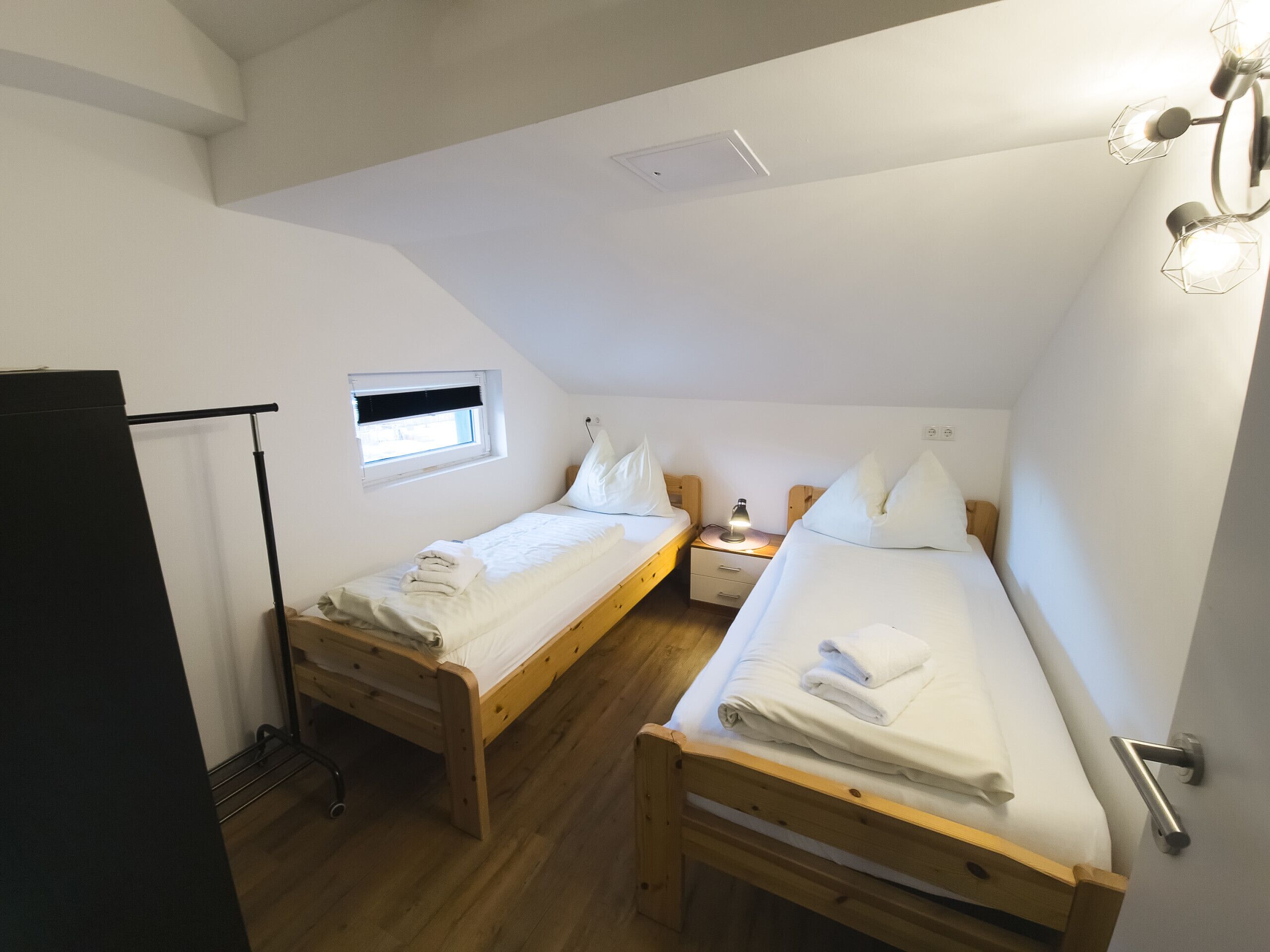 A small, well-lit room with two wooden beds, a window with blinds, a desk with a lamp, and a door.