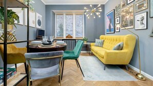 A modern living room with a yellow sofa, green chairs, a dining table with plates and glasses, and a large window with blinds. - Stylish Apartment with 2 Bedrooms | Close to the Center, SmartTV (Warszawa)
