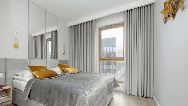A modern bedroom with a large bed, a bedside table, a mirror, and a window with curtains.