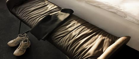 A bed with white linens, a brown padded bench, and a pair of white sneakers on the floor.
