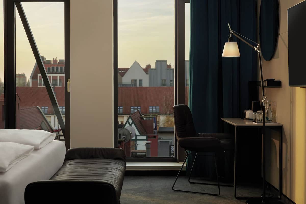 A hotel room with a bed, a desk with a lamp, a chair, and a view of buildings through large windows.