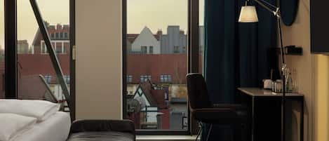 A hotel room with a bed, a desk with a lamp, a chair, and a view of buildings through large windows.