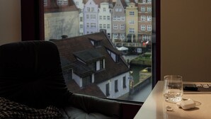 A room with a view of colorful buildings through a window, a leather chair, a glass of water, and a remote control on a table. - The Cloud One Gdansk (Gdansk)