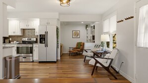 A modern kitchen with white cabinets, a built-in oven, and a refrigerator. There is a dining area with a table and chairs, and a small seating area with a chair and a plant.
