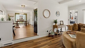 A spacious living room with a wooden floor, a sofa, a dining area with a table and chairs, and a kitchen in the background.