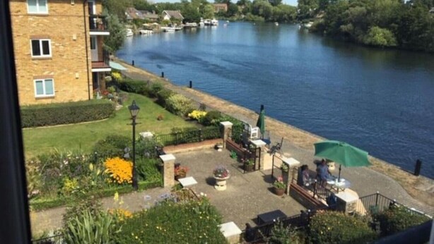 A riverside view with a building, a patio area with outdoor seating, and boats on the water. - Beautiful Penthouse Apartment (Shepperton)