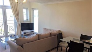 A living room with a beige sofa, a glass coffee table, a TV, and a dining area with a table and chairs. - Beautiful Penthouse Apartment (Shepperton)