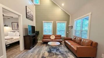 A living room with a sofa, a coffee table, and a TV. There are windows with blinds, a ceiling fan, and a door leading to another room.