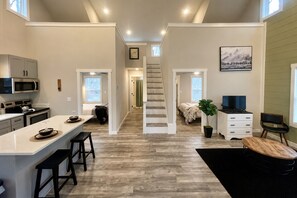 A modern kitchen with a central island, a dining area, a staircase leading to a bedroom, and a living area with a television and a plant.