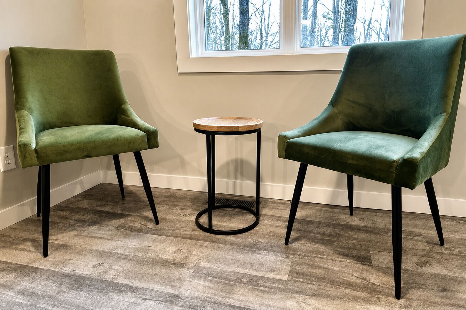 Kaksi vihreästi verhoiltua tuolia, joissa on mustat jalat, pieni pyöreä puupöytä ja ikkuna, josta avautuu näkymä paljaisiin puihin.