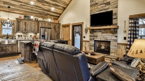 A spacious living area with a stone fireplace, leather sofa, and a kitchen with wooden cabinets and a stainless steel refrigerator.
