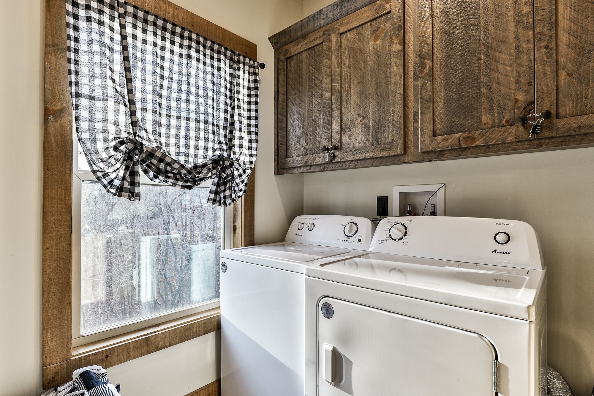 A laundry room with a white washer and dryer, wooden cabinets, and a window with a checkered curtain.
