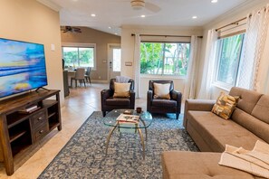A spacious living room with a large flat-screen TV, leather sofas, a glass coffee table, and a view of the outdoors through large windows. - 791 Sunset Cottage · NEW Heated Pool/Spa (Naples)
