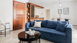 A living room with a blue sofa, a round coffee table with vases, a wooden cabinet, and a dining area in the background. - Faliro Sunlit 3BR Apartment (Palaio Faliro)