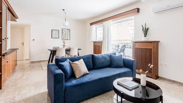 A modern living room with a blue sofa, a dining table with chairs, and a small side table with a book and vase. - Faliro Sunlit 3BR Apartment (Palaio Faliro)