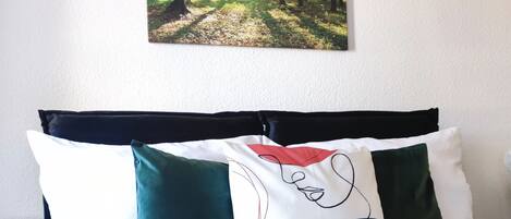 Una cama bien hecha con un edredón verde y varias almohadas, un cuadro de un bosque colgado en la pared y una mesita de noche pequeña.